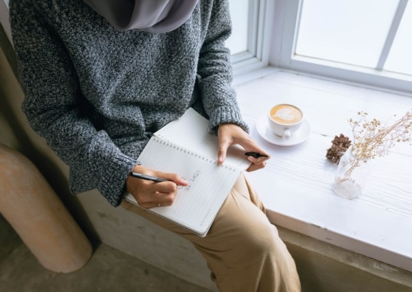 Frau sitzt am Fenster und schreibt in ein Buch als Beispiel für eine Selbstfürsorgeroutine im Alltag