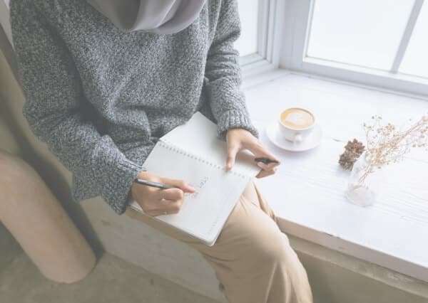 Frau sitzt am Fenster und schreibt in ein Buch als Beispiel für eine Selbstfürsorgeroutine im Alltag